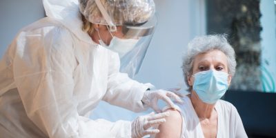 Profissional de saúde aplica vacina no braço de uma mulher idosa em ambiente interno.