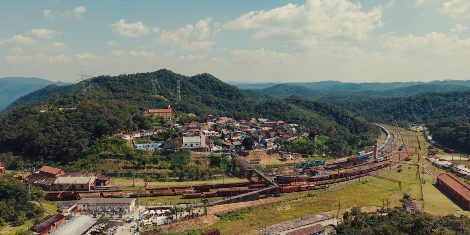 Vista aérea de uma pequena cidade entre morros verdes, atravessada por linhas ferroviárias.
