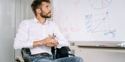 Homem em cadeira de rodas analisa gráficos em um quadro branco em um ambiente de escritório.