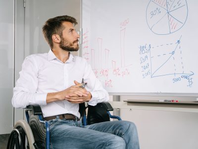 Homem em cadeira de rodas analisa gráficos em um quadro branco em um ambiente de escritório.
