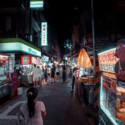 Feira noturna ao ar livre, com barracas de comida iluminadas e pessoas circulando entre os estandes.