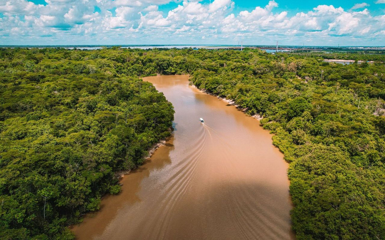 Vista aérea de uma embarcação navegando por um rio barrento cercado pela floresta amazônica.