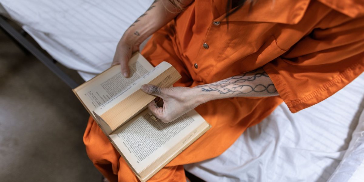 Mulher privada de liberdade lê um livro sentada em uma cama dentro de uma cela.
