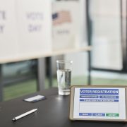 Tablet exibe um formulário digital de registro eleitoral em uma seção de votação, com placas indicando o dia da eleição.