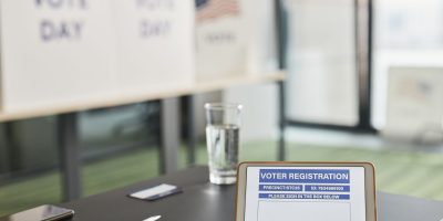 Tablet exibe um formulário digital de registro eleitoral em uma seção de votação, com placas indicando o dia da eleição.