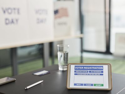 Tablet exibe um formulário digital de registro eleitoral em uma seção de votação, com placas indicando o dia da eleição.